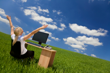 Business concept shot of a beautiful young woman sitting at a desk using a computer in a green field raising her arms into a bright blue sky with fluffy white clouds. Shot on location with copyspace at the top of the image.
